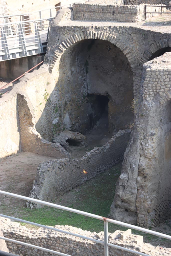 III.1/2/18/19, Herculaneum, October 2023.
Looking north-west from access roadway towards remains of oven/hearth in room G.
Photo courtesy of Klaus Heese.
According to Pagano –
“Nelle altre localita che restano dall’altro lato della strade, (….. da Ins.II/1…..), allo stesso livello di detti magazzini, eravi una officina di panizzazione, argomentandosi da un forno ivi trovato, di cui resta qualche avanzo, e pel rinvenimento di diversi grandi vasi in terracotta che contenevano farina”.
(In another place that remains on the other side of the roadway, (…..from Ins. II/1…..), and at the same level of these warehouses, there was a bakery, arguably from an oven found there, of which remains some leftovers, and from the discovery of several large terracotta pots which contained flour.)
See Pagano, N. Descrizione degli Scavi di Ercolano, 1870. (p.29).
According to Maiuri, he thought room G would appear to be the services (kitchen) for the two large sitting rooms (oeci) –
“Gli ambienti rustici (G-M) con cui si chiede a ponente il quartiere sotterraneo, semidistrutti e interrati, sono per loro stessa disposizione e orientazione in servizio dei due grandi oeci di soggiorno e di convito: nell’ambiente G resta la traccia di un focolare; in corrispondenza dell’ambiente I alcuni gradini (h) accennano alla presenza di un cellaio o repositorio sottostante.”
(The rustic rooms (G – M), which close off the underground rooms on the west side, semi-destroyed and buried, are by their very arrangement and orientation in service of the two large oeci for rest and dining: in room G the trace of a hearth remains; in room I some steps (h) hint at the presence of a cellar or storage room below.)
See Maiuri, A., 1958. Ercolano, I Nuovi Scavi (1927-1958). (p.334).
